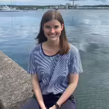 Frau am Wasser sitzend, lächelt in die Kamera.